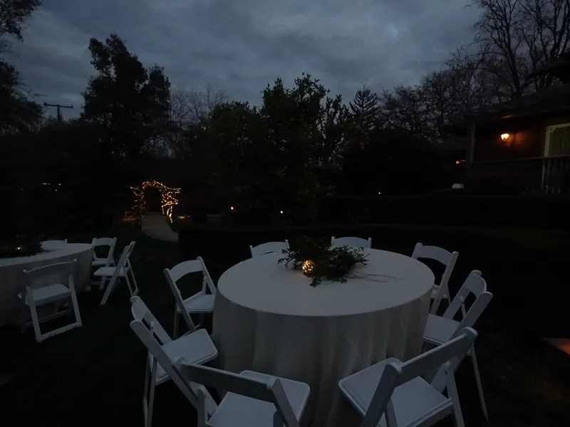 Round guest table with white folding chairs set in a backyard at twilight