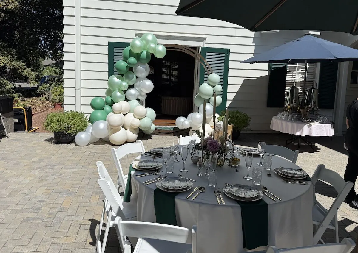 Sage and ivory balloon garland framing a doorway above a table styled with linens and fresh stems — balloon and bloom styling for San Ramon catering