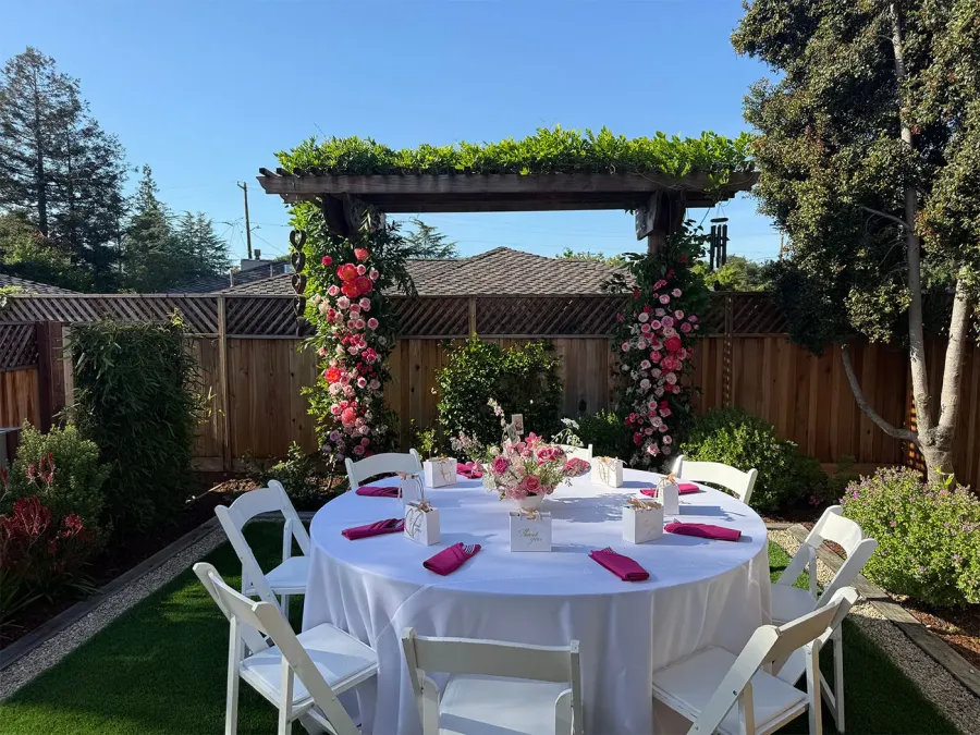 Rose arch in pink and red curving over a round garden table laid with white linens and fuchsia napkins — custom arch installations for Pleasanton catering events